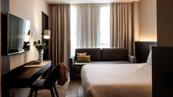 Une chambre d’hôtel comprenant un lit, un canapé, un bureau avec une chaise, un téléviseur à écran plat et une grande fenêtre avec des rideaux.