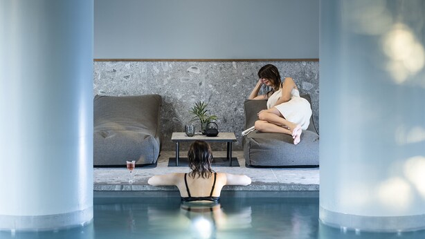 A modern indoor pool area with a clear view of a person sitting on a couch, a small table with a plant, and a glass of water. - Litta Palace Milano Urban Resort (Lainate)