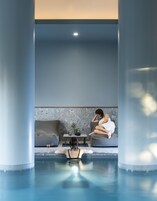 Un área de piscina interior moderna con vista despejada a una persona sentada en un sofá, una mesita con una planta y un vaso de agua.