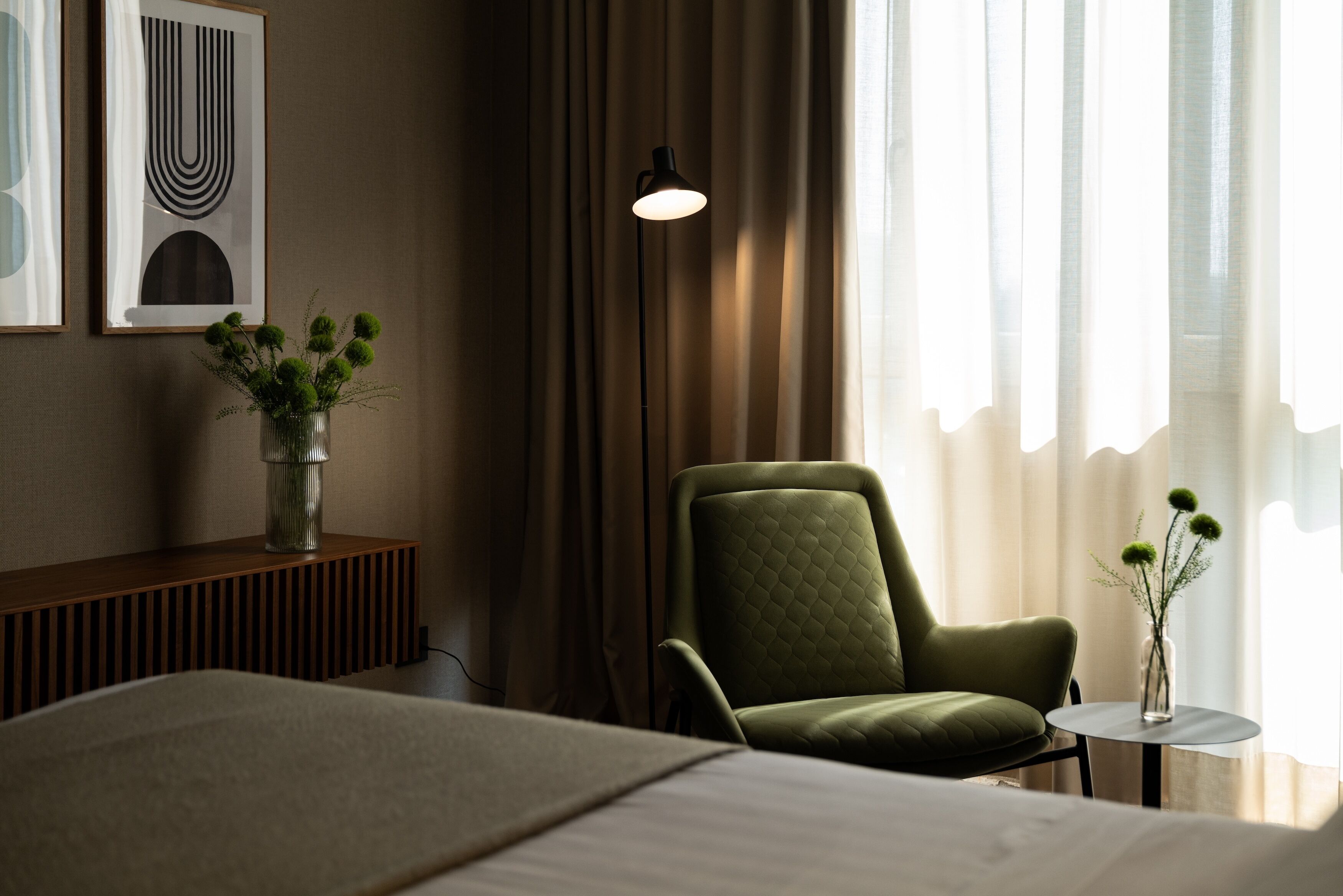 Une chambre d’hôtel avec un lit, un fauteuil vert, une petite table ronde ornée d’un vase de fleurs et une lampe fixée au mur.
