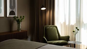 Une chambre d’hôtel avec un lit, un fauteuil vert, une petite table ronde ornée d’un vase de fleurs et une lampe fixée au mur.