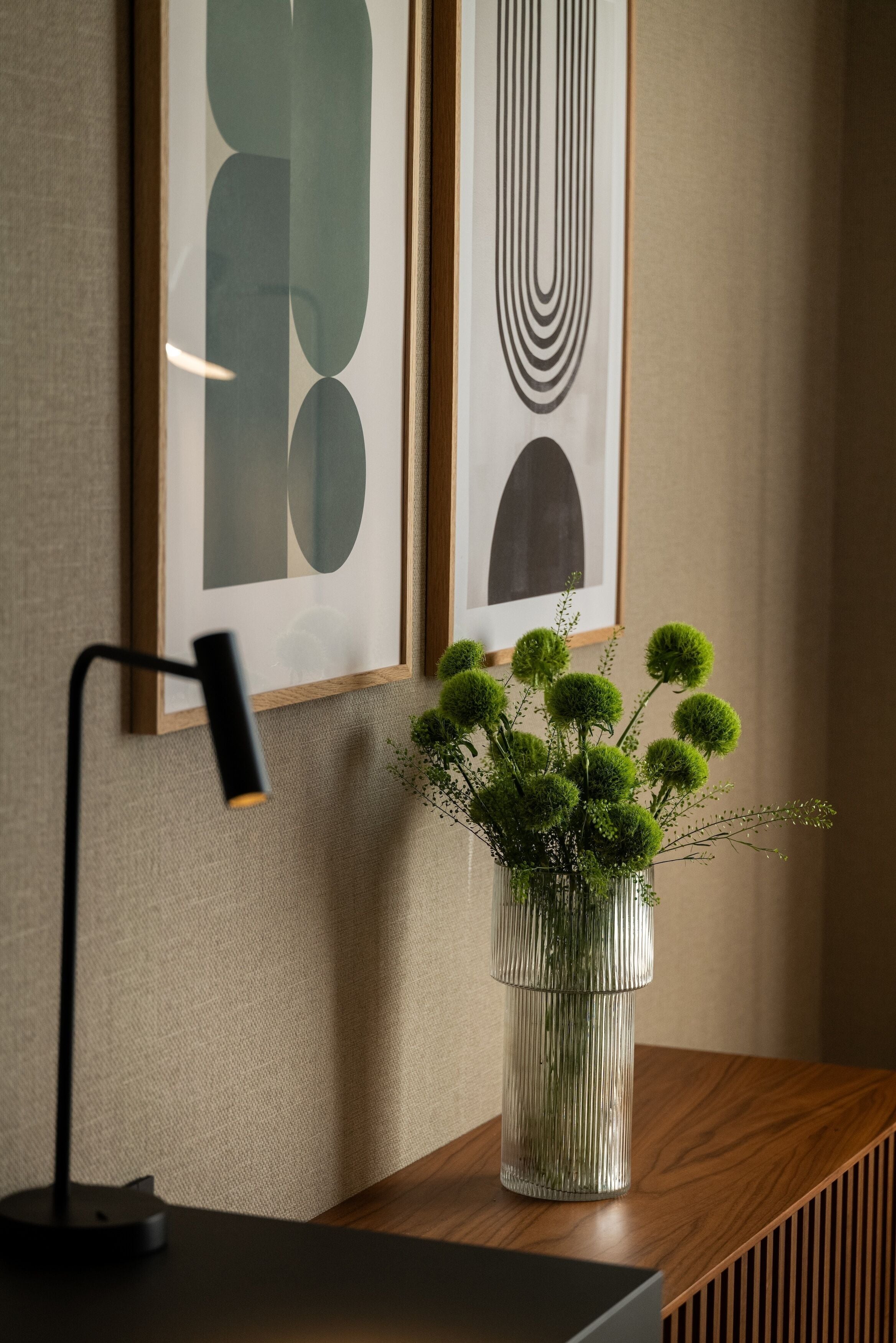Ein gerahmtes abstraktes Kunstwerk an der Wand, eine moderne Schreibtischlampe und eine Vase mit kugelförmigen grünen Blumen.