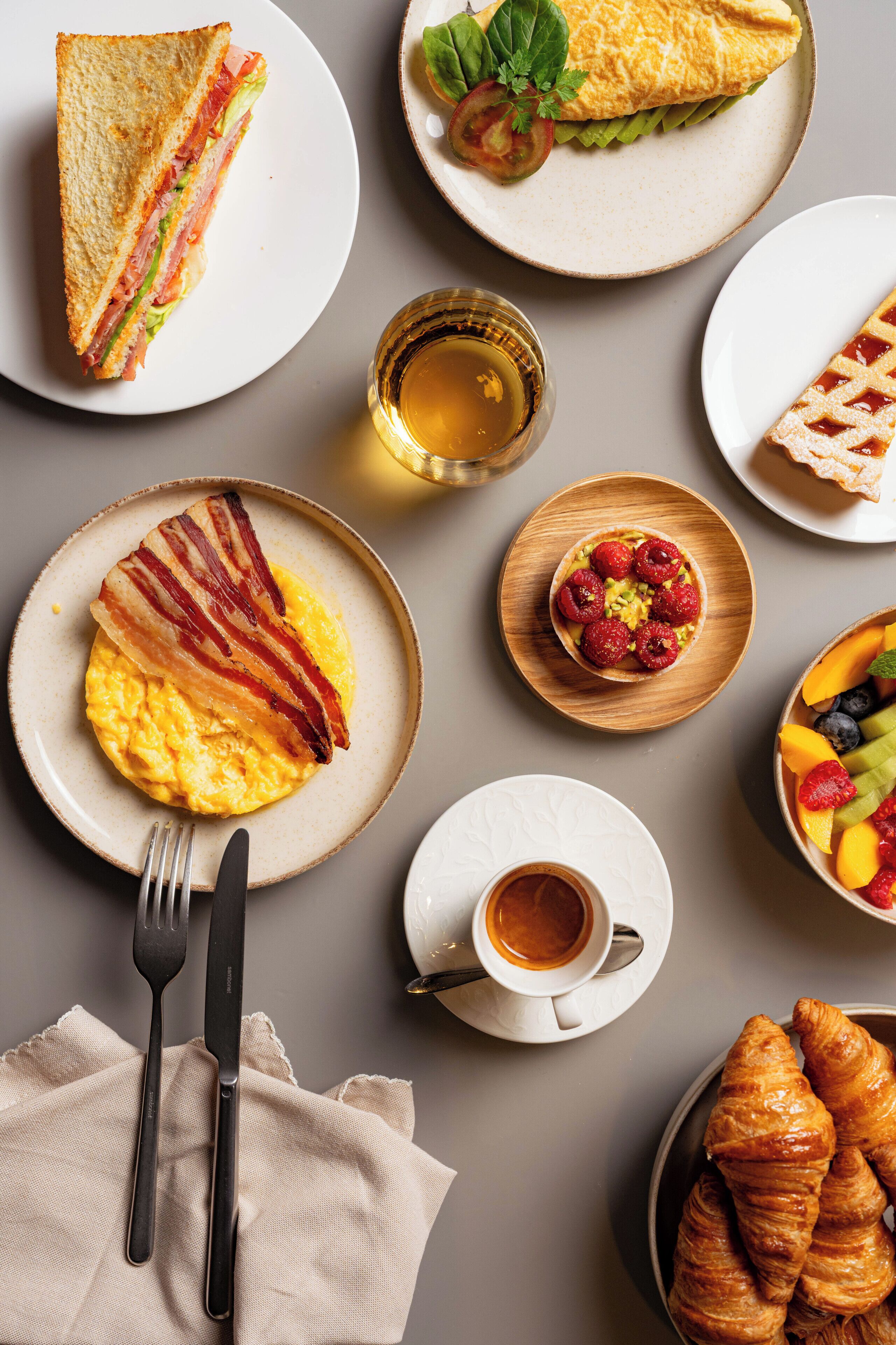 A breakfast setting with a sandwich, omelette, croissants, and a fruit bowl.