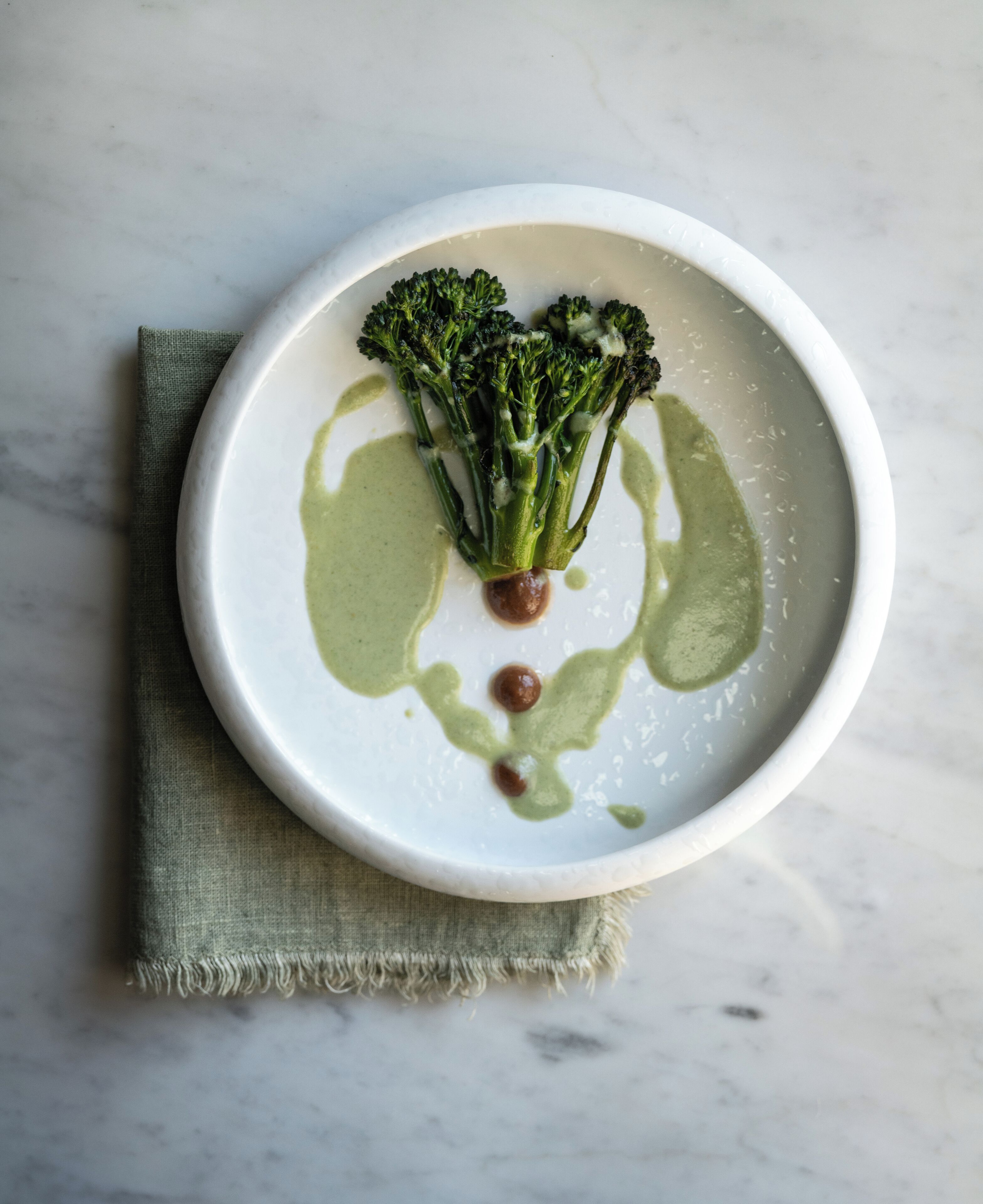 Une assiette blanche sur laquelle des bouquets de brocolis sont disposés de manière à former un visage, accompagnée d’une sauce verte et d’un petit élément brun, le tout posé sur une surface en marbre recouverte d’un tissu vert.