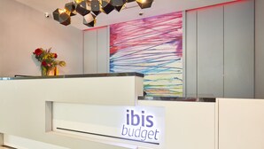 Een receptiebalie met een bordje dat 'ibis budget' vermeldt, een kleurrijk abstract schilderij aan de muur en een vaas met rode bloemen.
