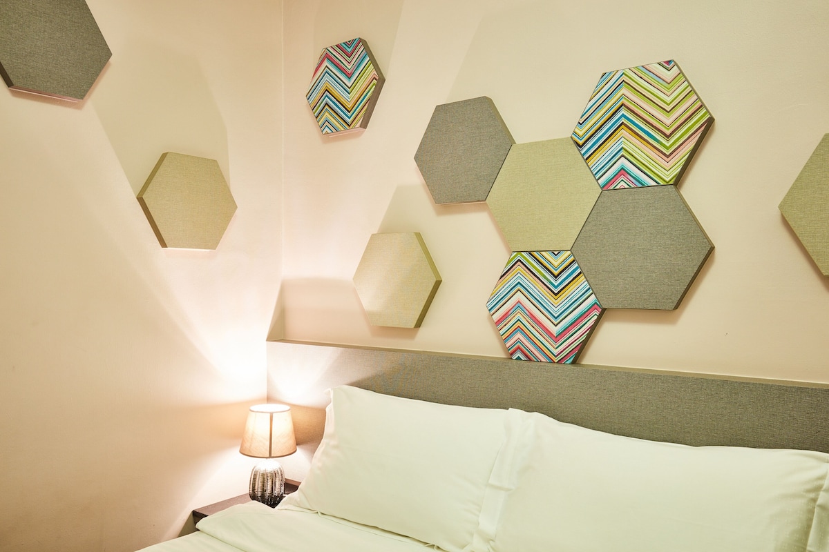 Una habitación de hotel con una cama, una mesita de noche, una lámpara y un muro decorado con espejos hexagonales.