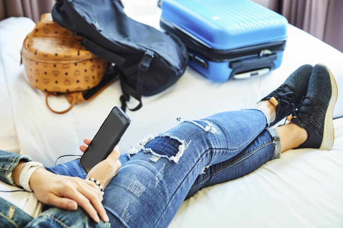 Uma pessoa deitada na cama com um celular, uma mochila preta e uma mala azul.