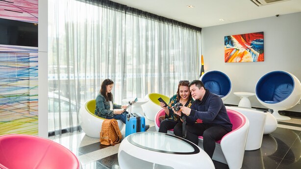 Een moderne hotellobby met kleurrijke stoelen, een groot raam met lichte gordijnen en een schilderij aan de muur.