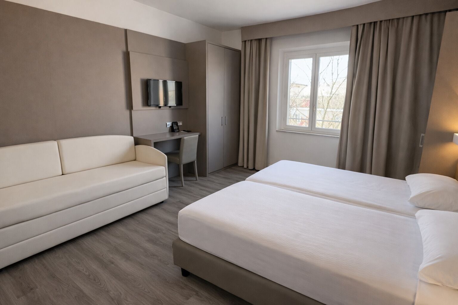 Habitación de hotel con cama, sofá, escritorio, televisor y ventana con cortinas.