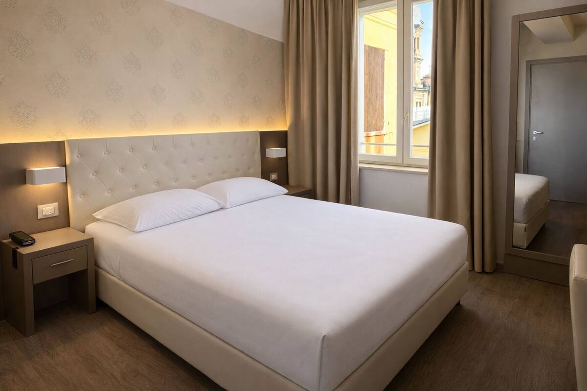 Una cama bien hecha con cabecera, una mesita de noche con lámpara y una ventana con cortinas.