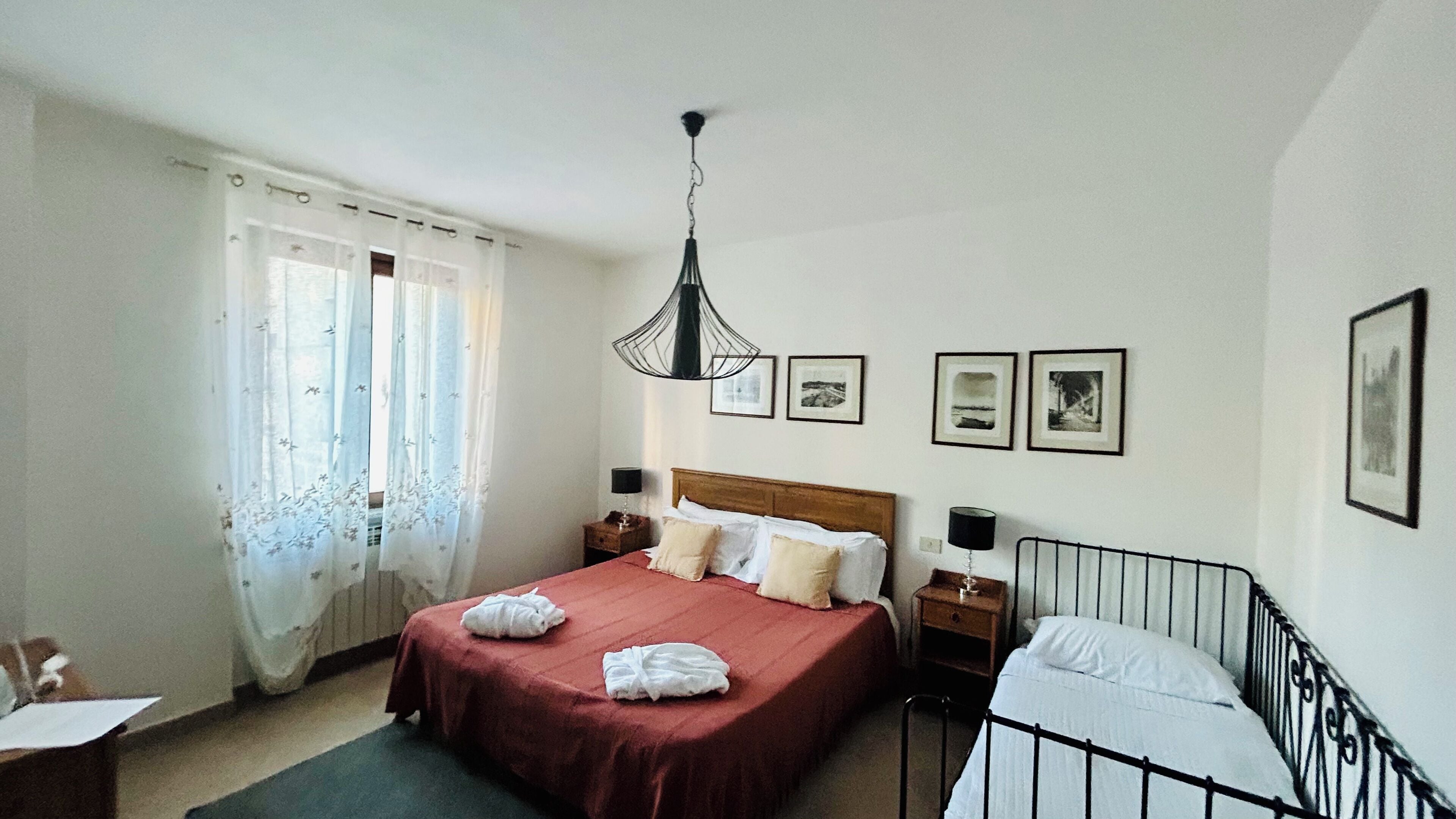 Un dormitorio con una cama grande, una mesita de noche, una lámpara colgante y una ventana con cortinas.
