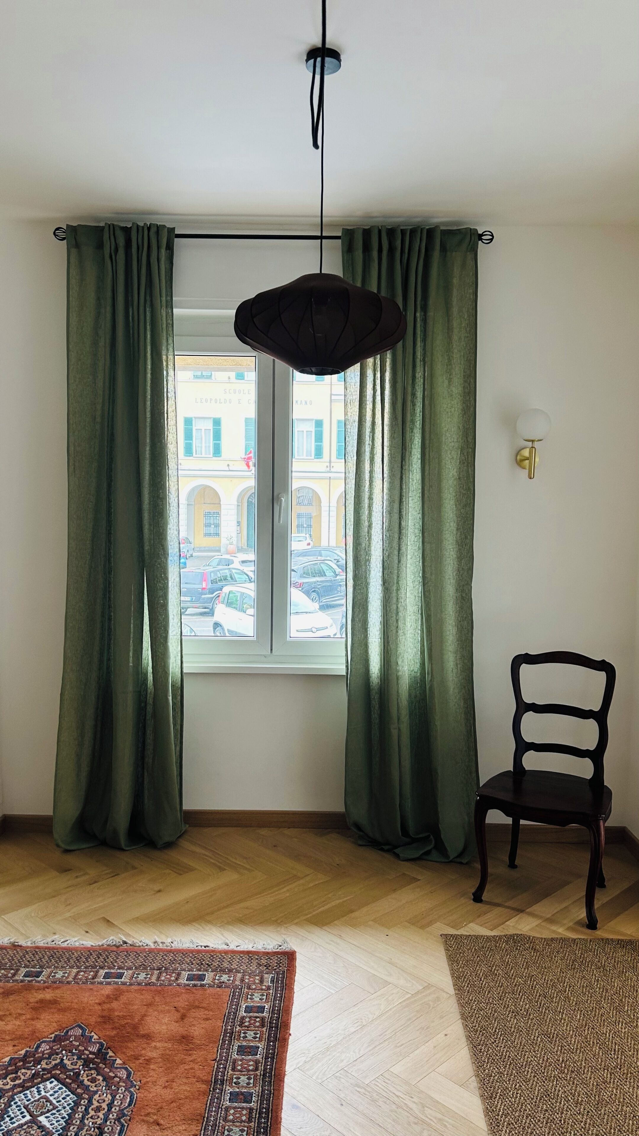 Una stanza con finestra, tende, una sedia, un lampadario e un tappeto decorato.