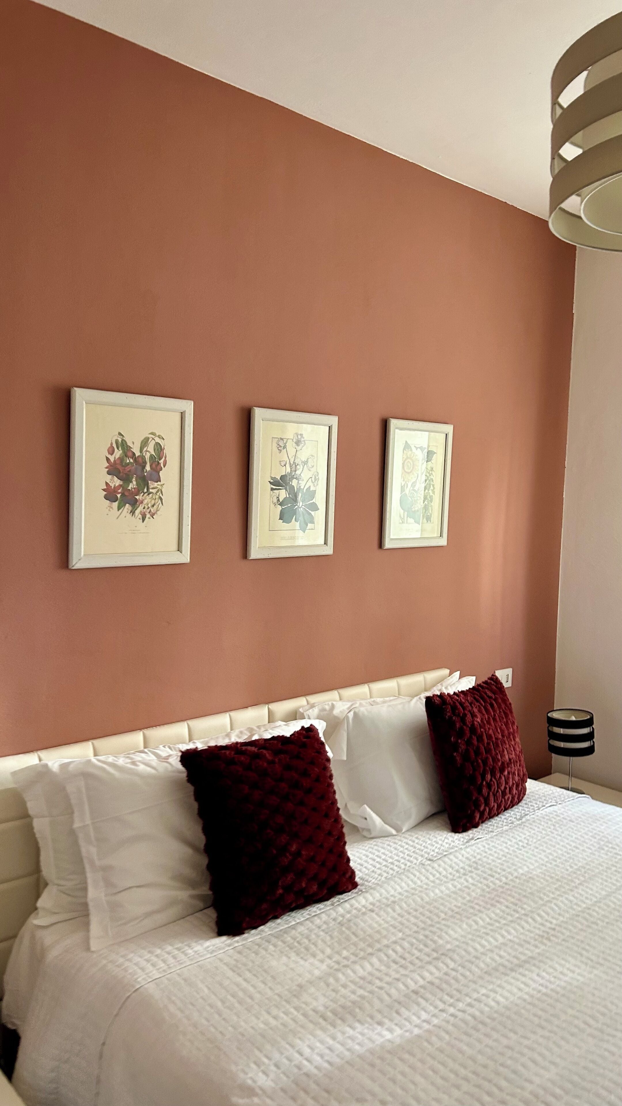 Habitación de hotel con cama, ropa de cama blanca, dos almohadas rojas y tres láminas botánicas enmarcadas en la pared.