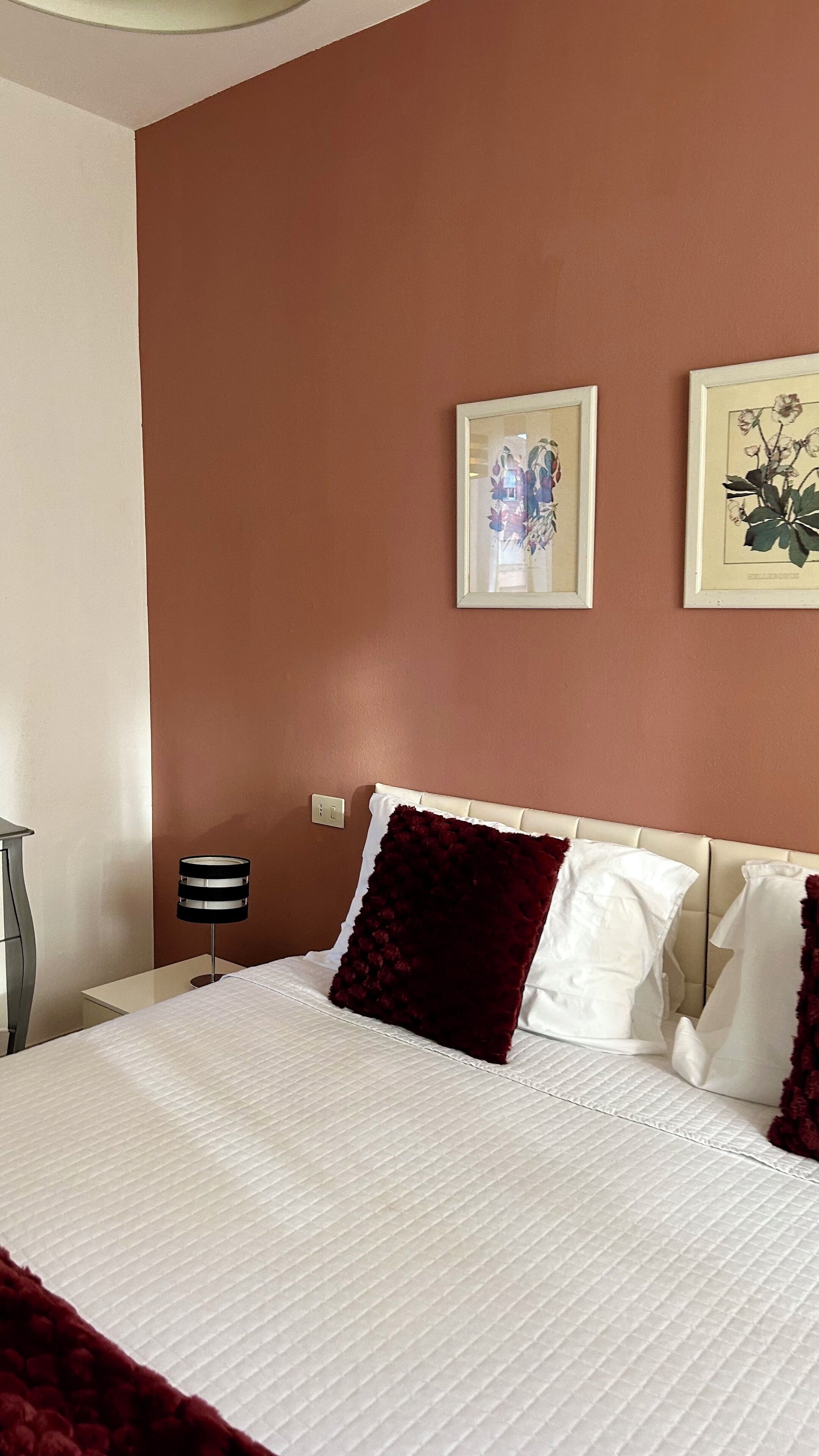 A neatly made bed with white linens and a red accent pillow, a bedside lamp, and two framed botanical prints on the wall.