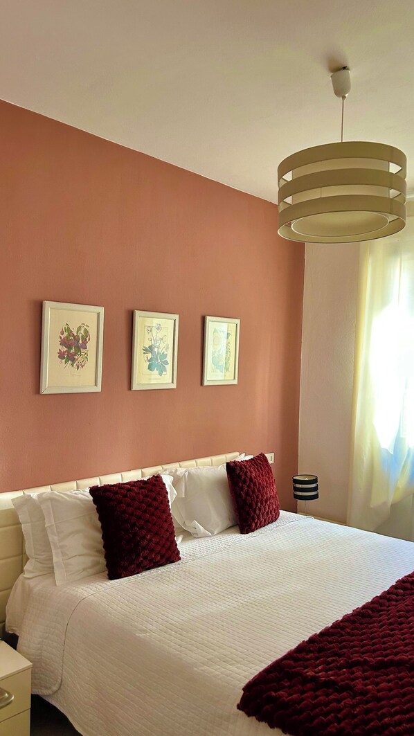 Un dormitorio con una cama, dos láminas botánicas enmarcadas en la pared, una lámpara de techo y una mesita de noche con lámpara.