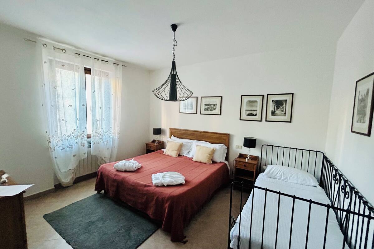 A bedroom with a large bed, a bedside table, a nightstand, a hanging light fixture, and a window with curtains.