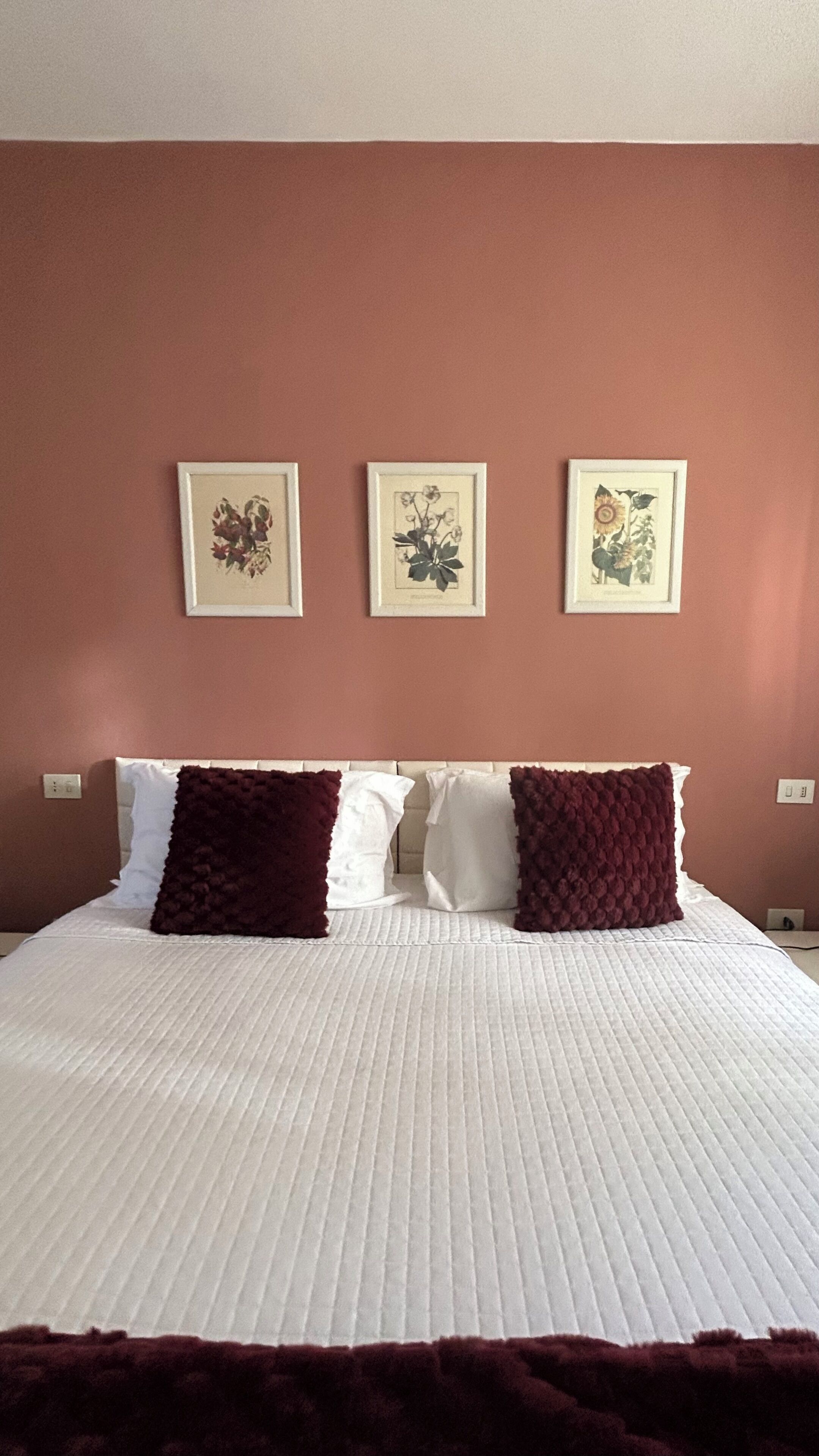 A neatly made bed with white bedding and two dark red pillows, set against a wall with three framed floral artworks.