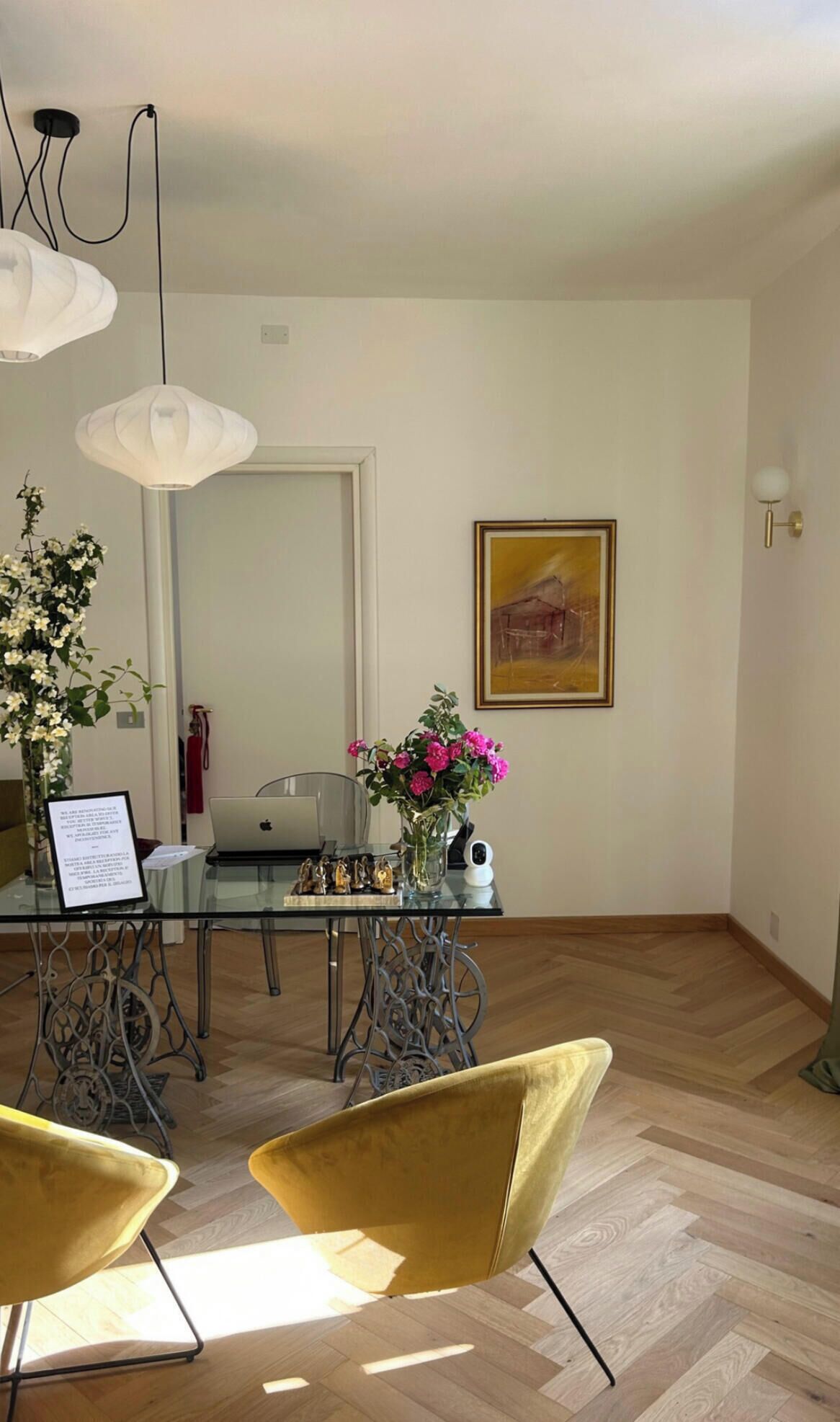 A dining area with a glass table, metal chairs, a laptop, and a vase with flowers.