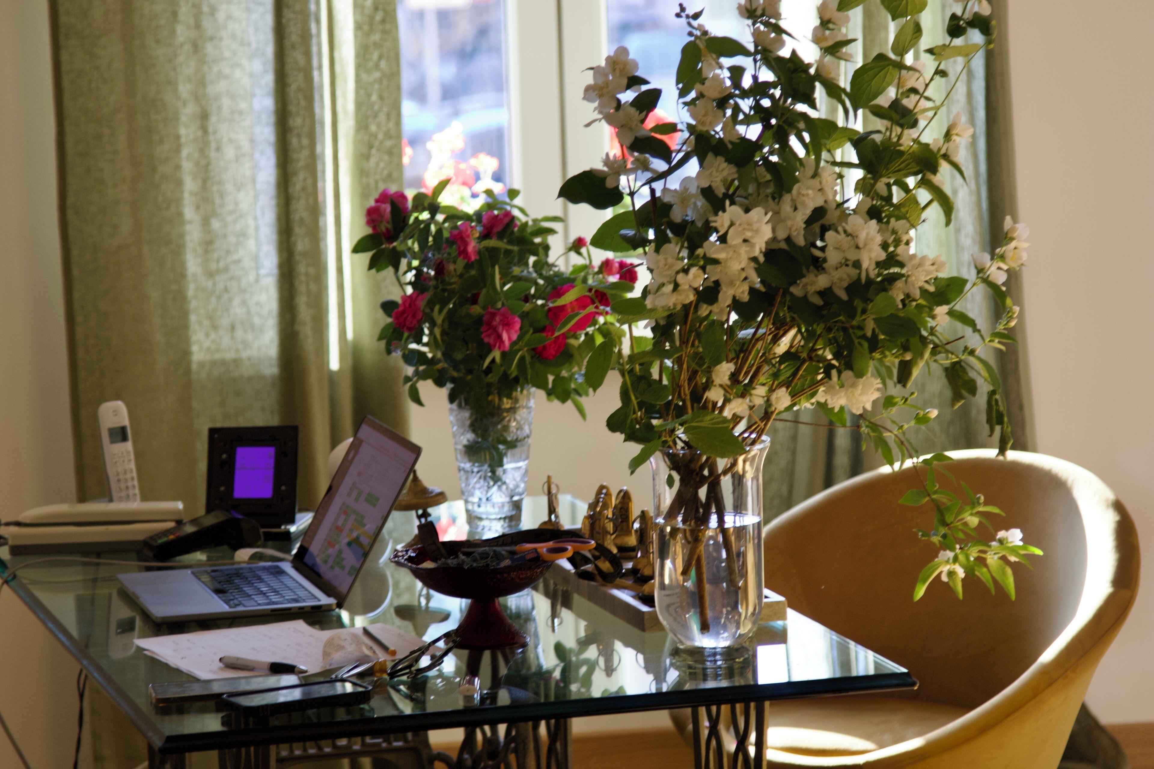Ein Glastisch mit einem Laptop, einer Blumenvase, einem Stuhl und einem Fenster mit Vorhängen.