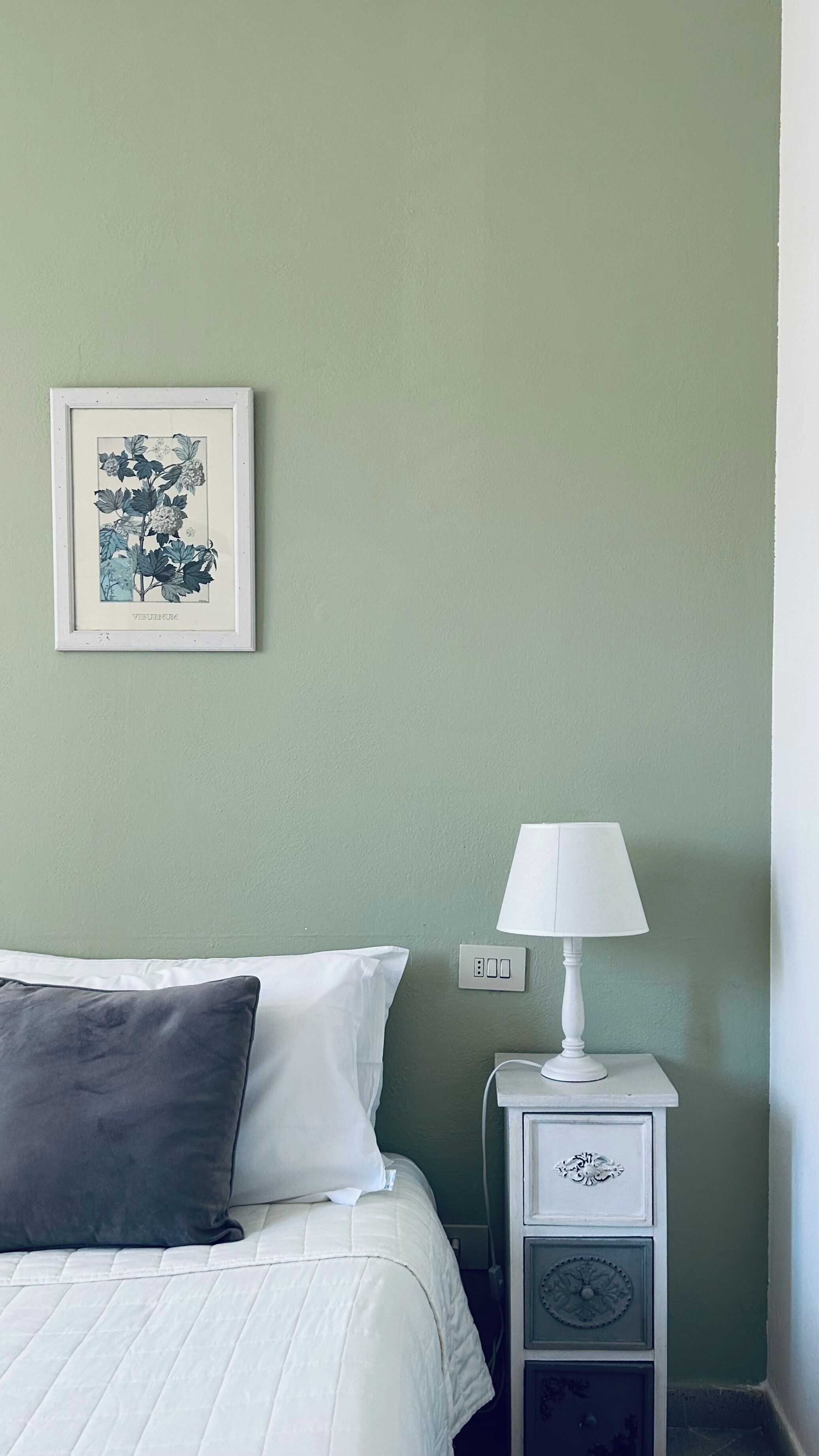 Una cama bien hecha con almohadas blancas y grises, un grabado botánico enmarcado en la pared, una mesita de noche blanca con una lámpara y un enchufe.