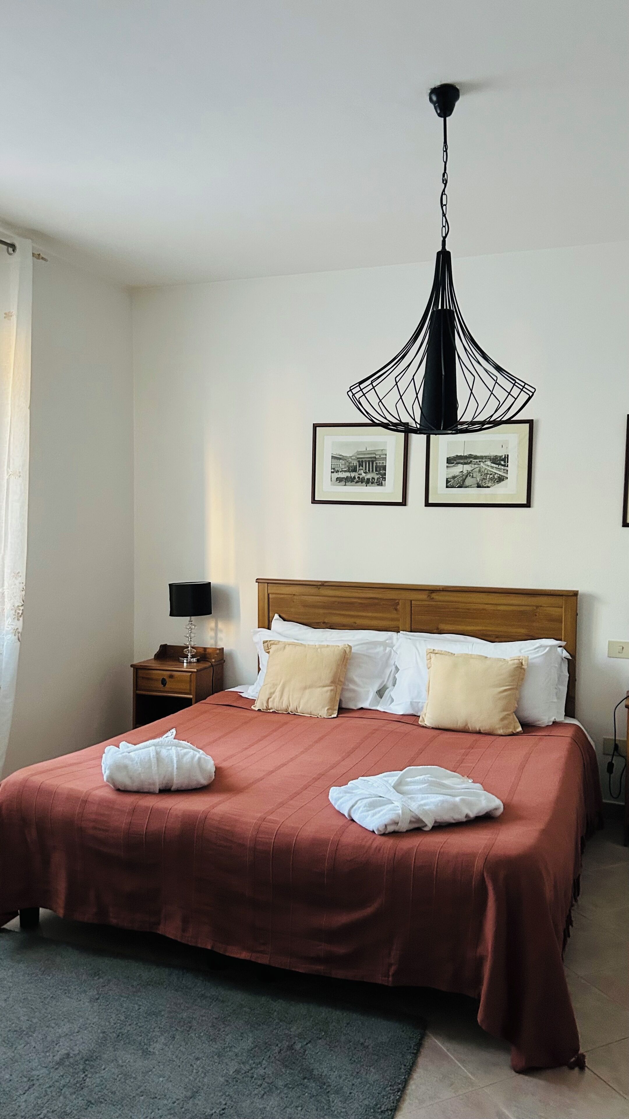 A neatly made bed with a red bedspread and white pillows, a wooden nightstand with a lamp, and a black hanging light fixture above.