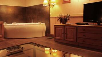 A modern bathroom with a large bathtub, wooden cabinets, a flat-screen TV, and a glass coffee table with decorative items.