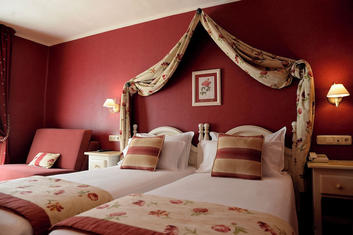 Habitación de hotel con dos camas, una pared roja, cortinas con motivos florales y lámparas de noche.