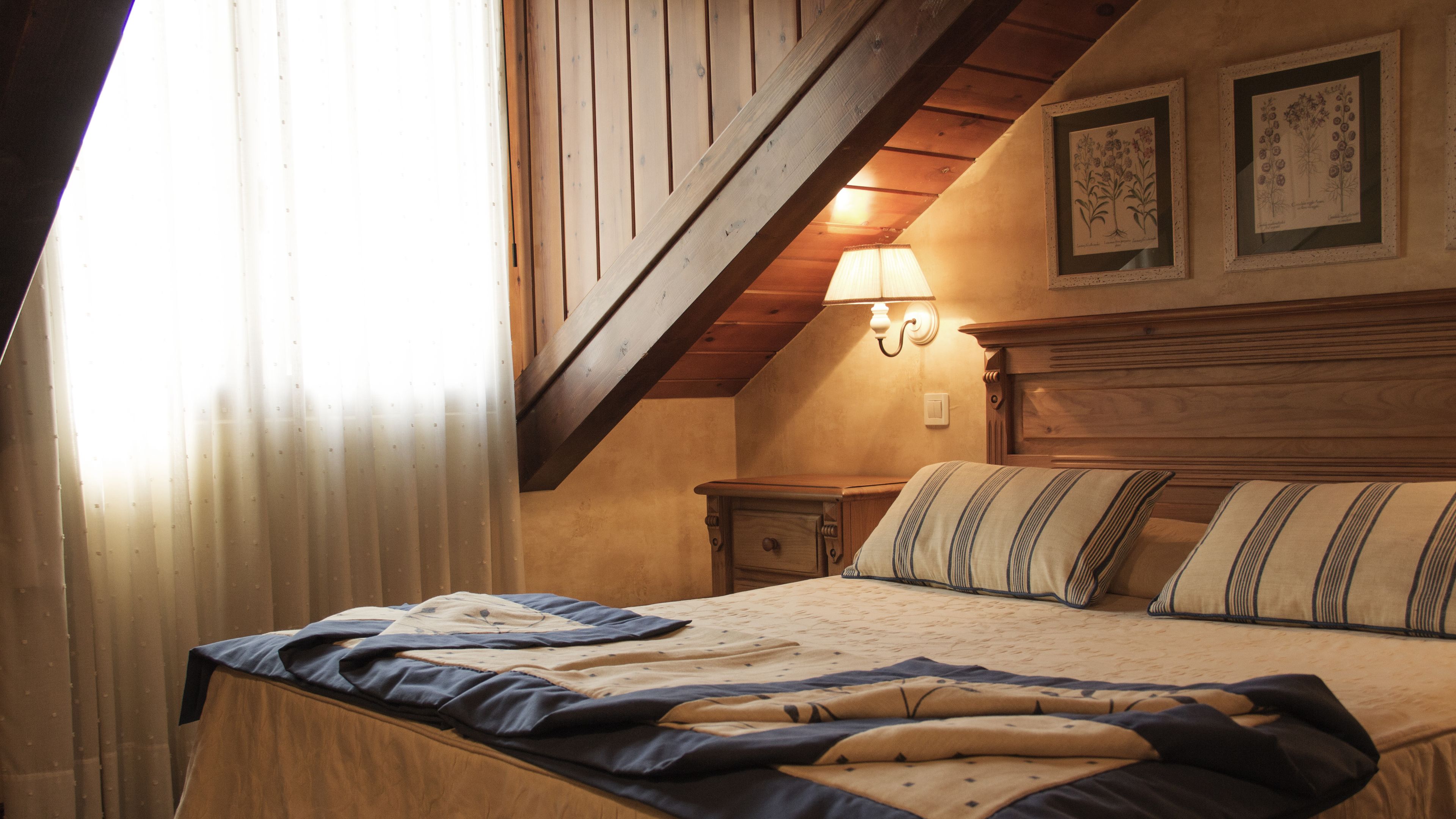 Un dormitorio con techo de madera, una cama con ropa de cama a rayas, una mesita de noche e impresiones botánicas enmarcadas en la pared.