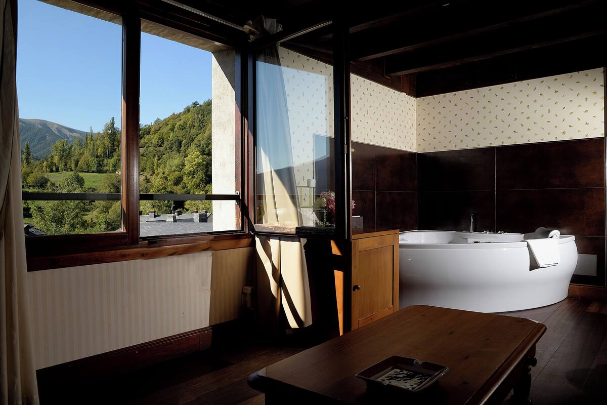 Un baño con una bañera grande, un banco de madera y vistas a un paisaje verde a través de una ventana.