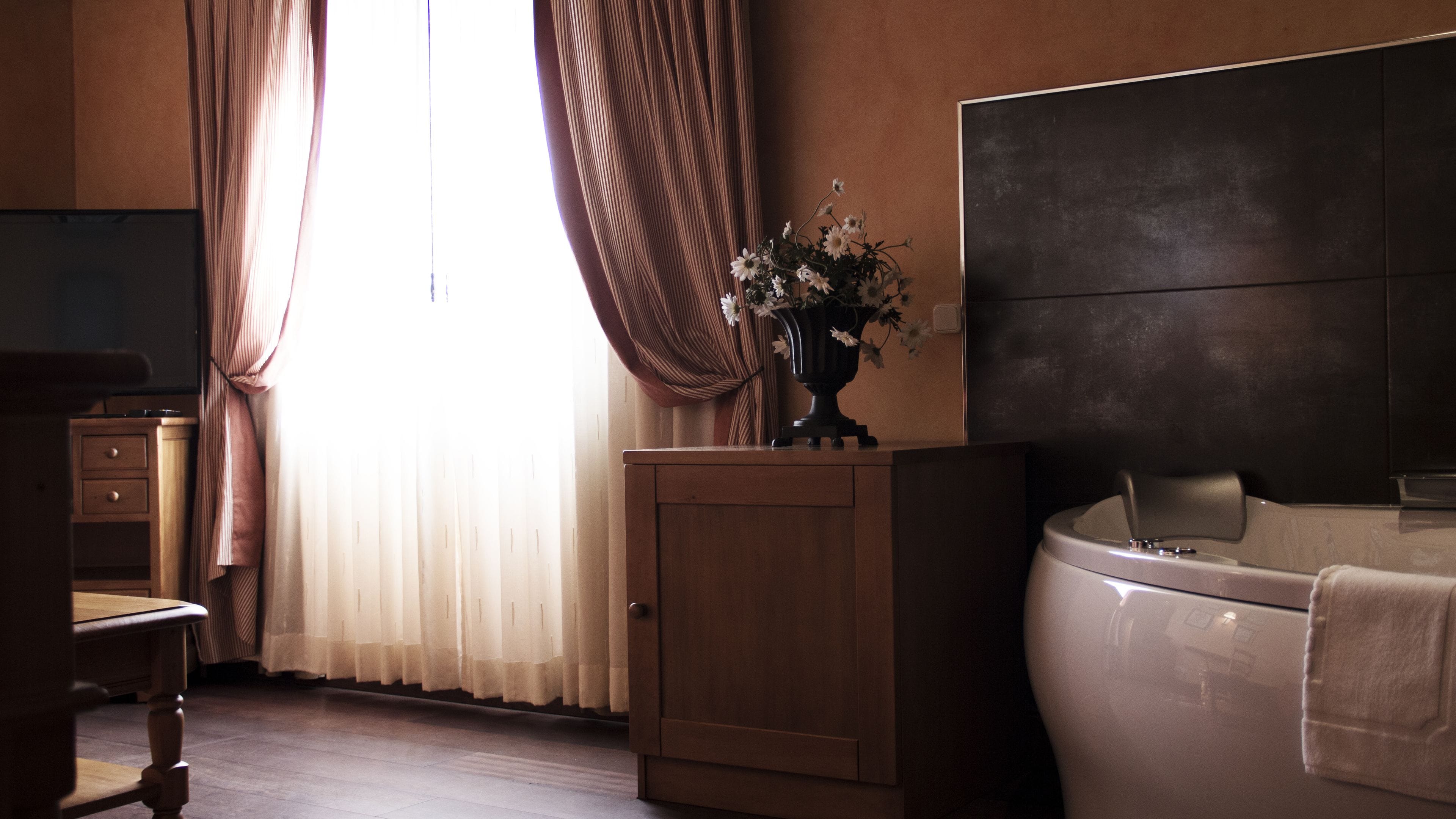 A hotel room with a wooden cabinet, a vase with flowers, a large bed, and a window with curtains.