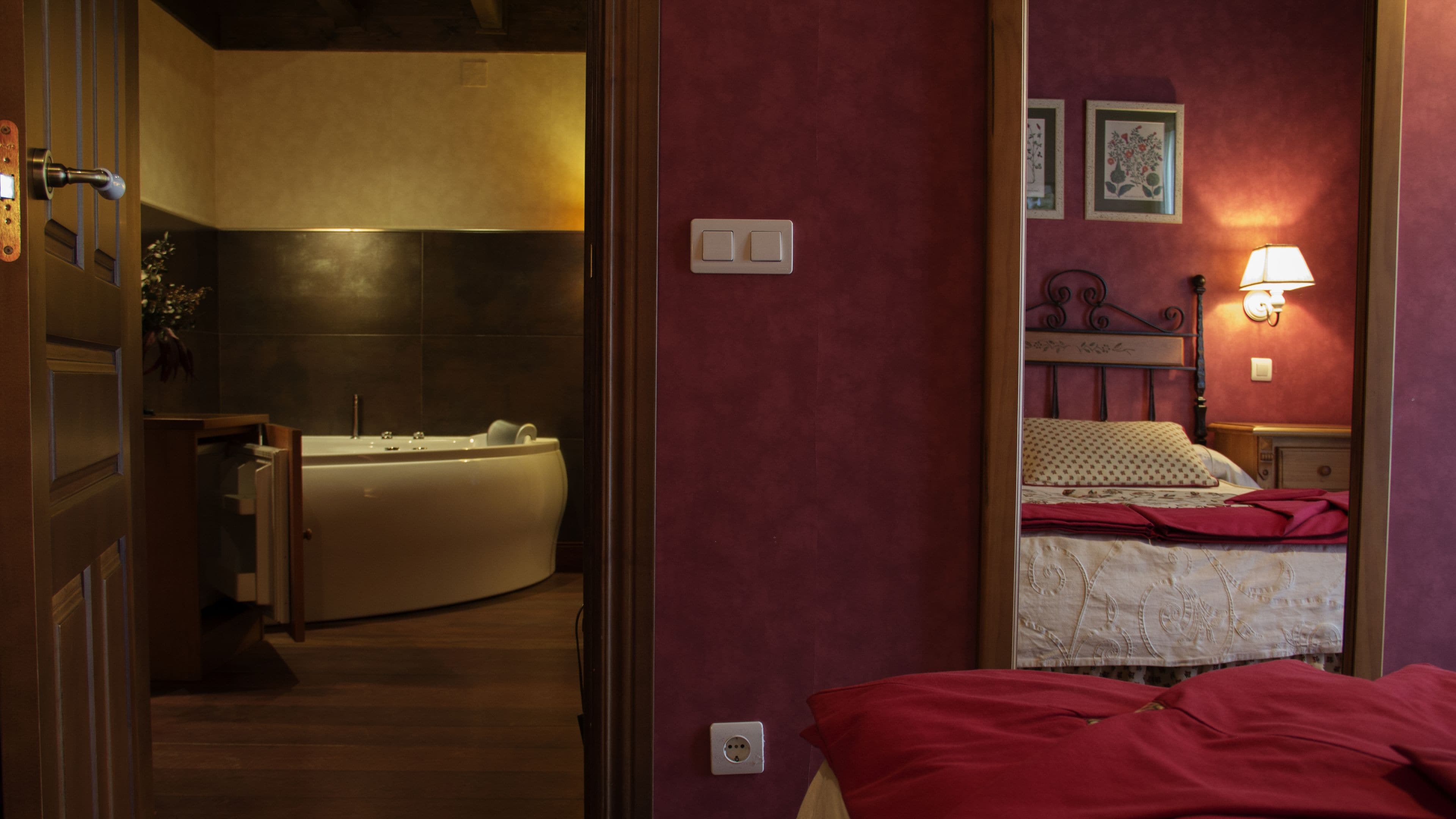 A hotel room with a bathtub, a bed with a red blanket, and a mirror reflecting the room.
