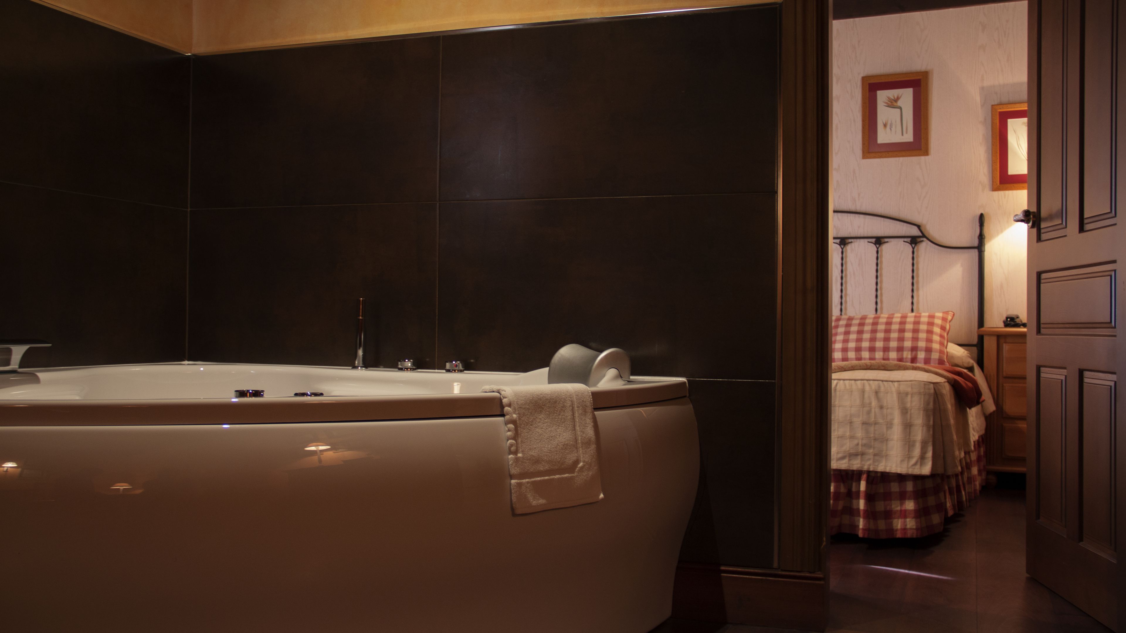 A modern bathroom with a large bathtub, dark tiles, and a visible door leading to a bedroom with a checkered bedspread.