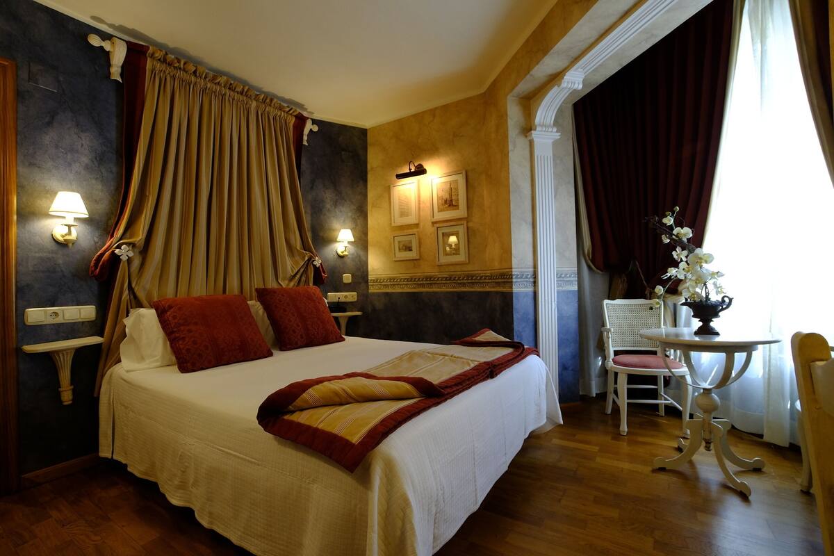 Habitación de hotel con una cama grande, una mesita de noche con lámpara, una silla y una pequeña mesa con un florero.