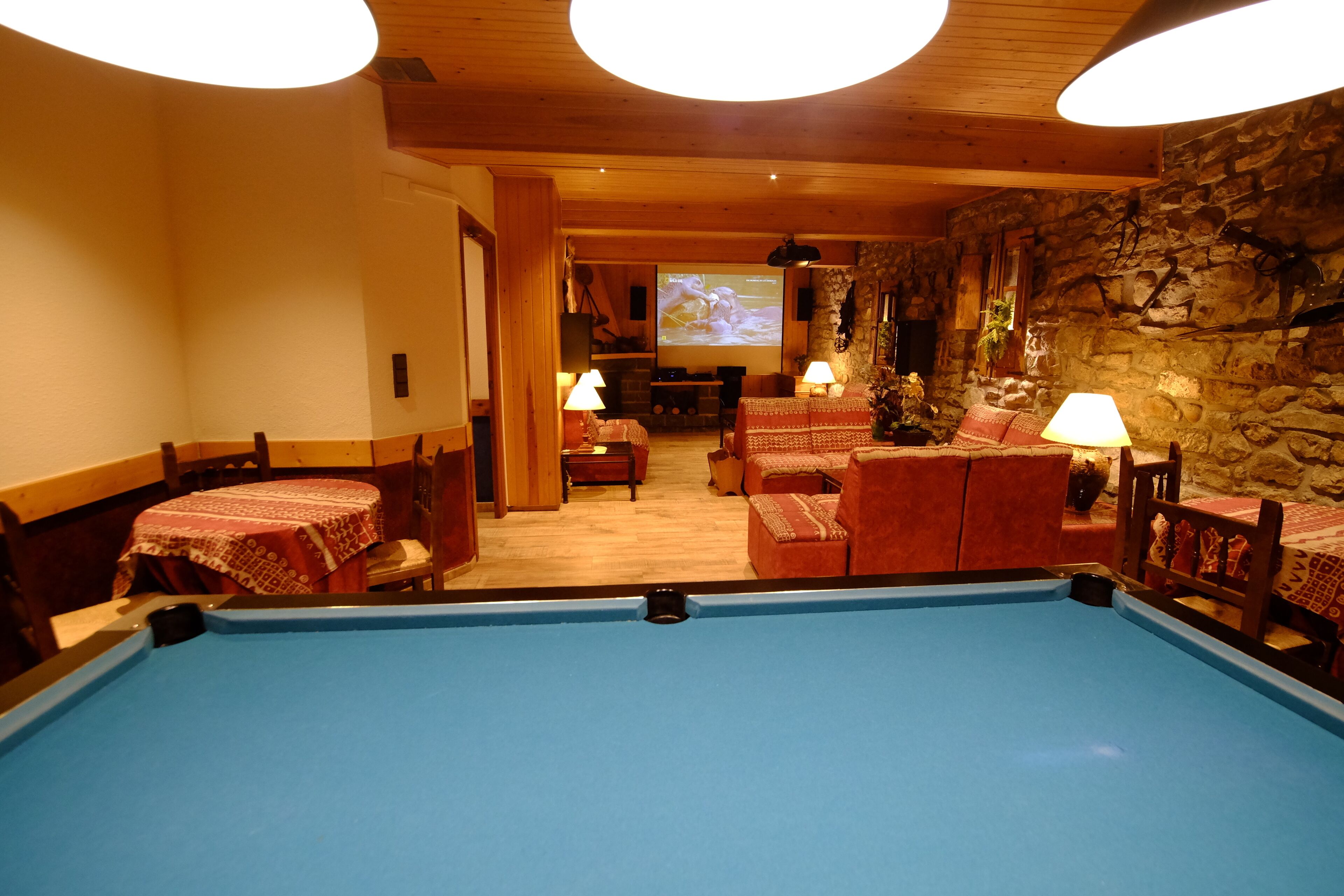 A pool table in a room with wooden walls and stone accents, furnished with red upholstered chairs and tables, and a fireplace.