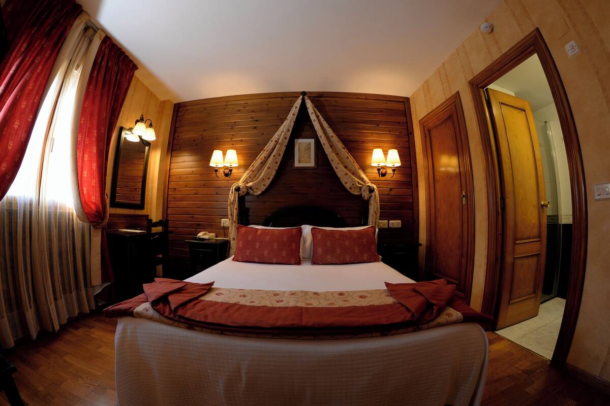 Habitación de hotel con techo curvo, cama con ropa de cama roja, dos lámparas fijadas a la pared y una puerta de madera.