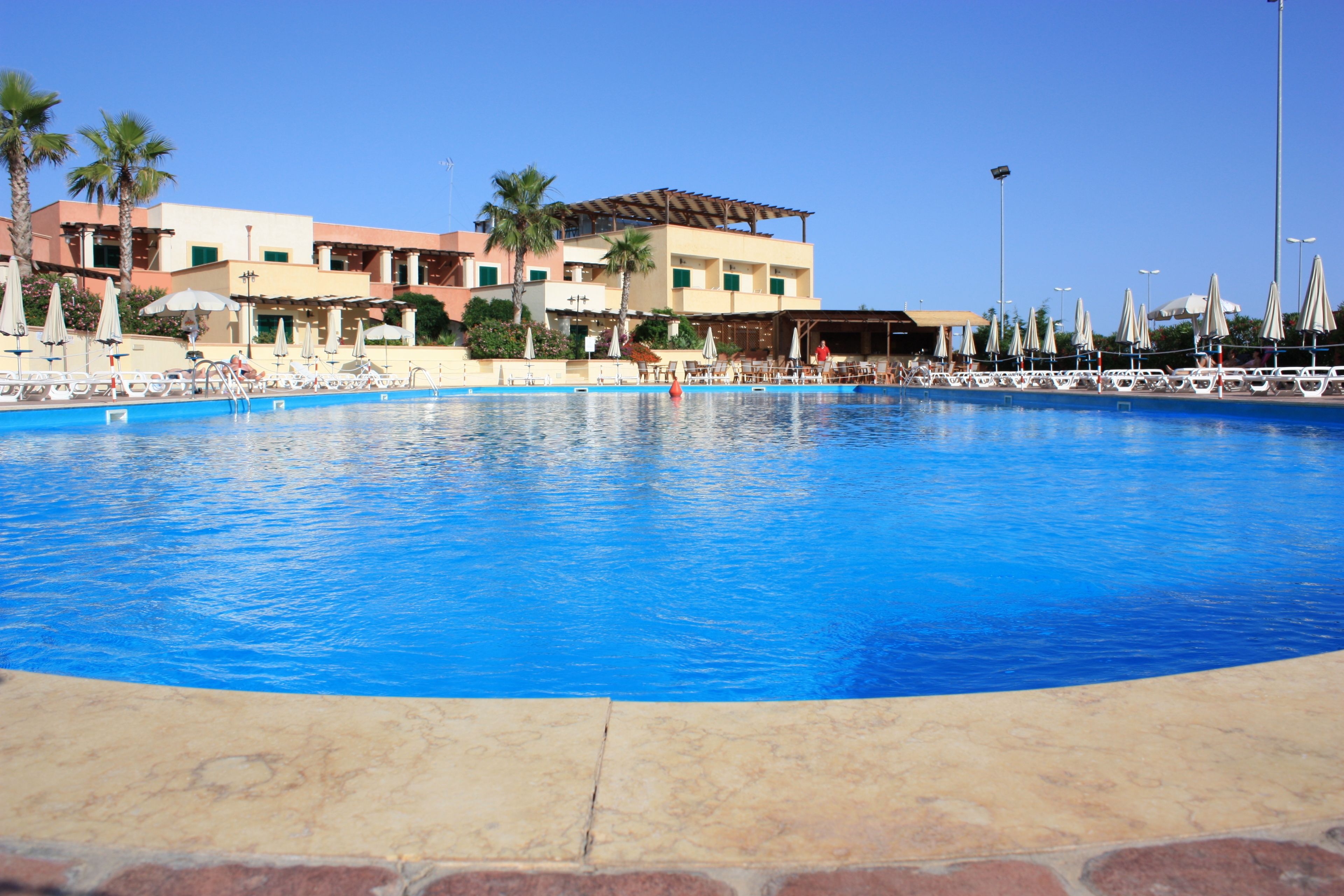 A large swimming pool with lounge chairs and umbrellas, surrounded by buildings with palm trees and a clear sky.