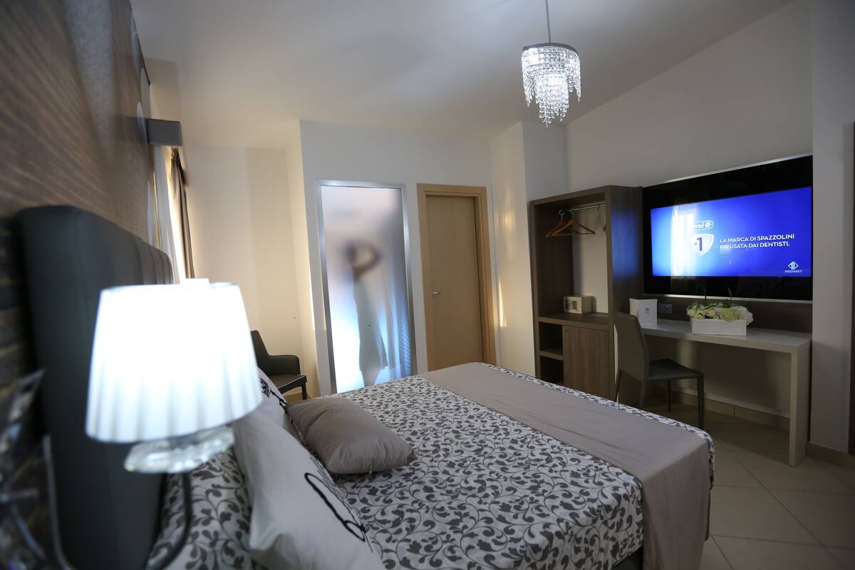 A modern hotel room with a bed, a desk, a television, and a chandelier.