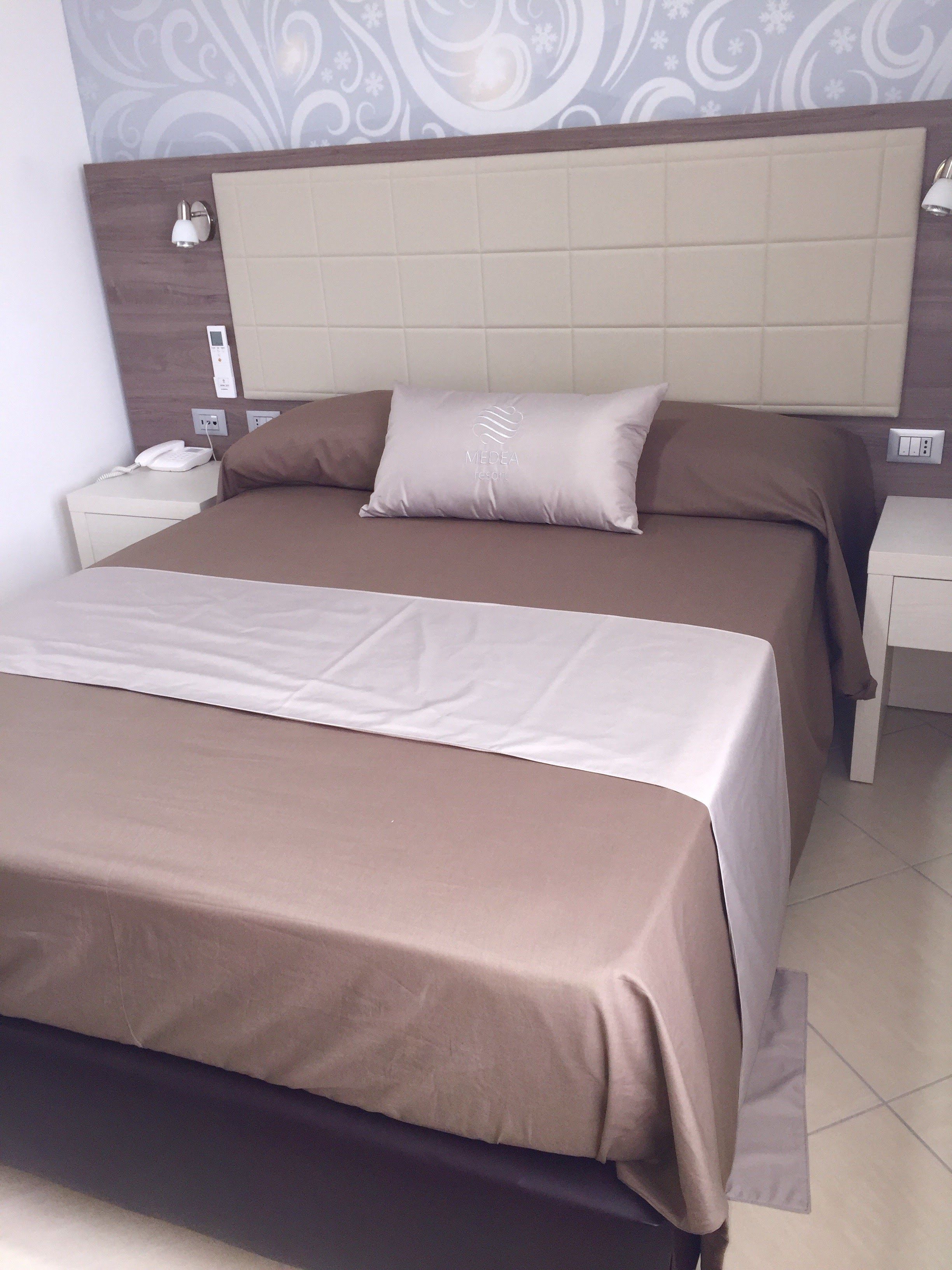 Una cama bien hecha con una colcha marrón y almohadas, un teléfono en la mesita de noche y una lámpara de pared.