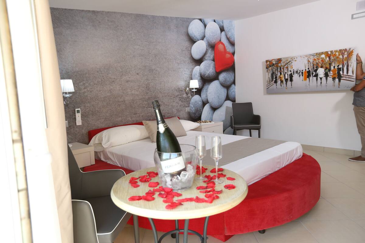 A modern hotel room with a bed, a small table with a bottle of champagne and glasses, and a painting on the wall.