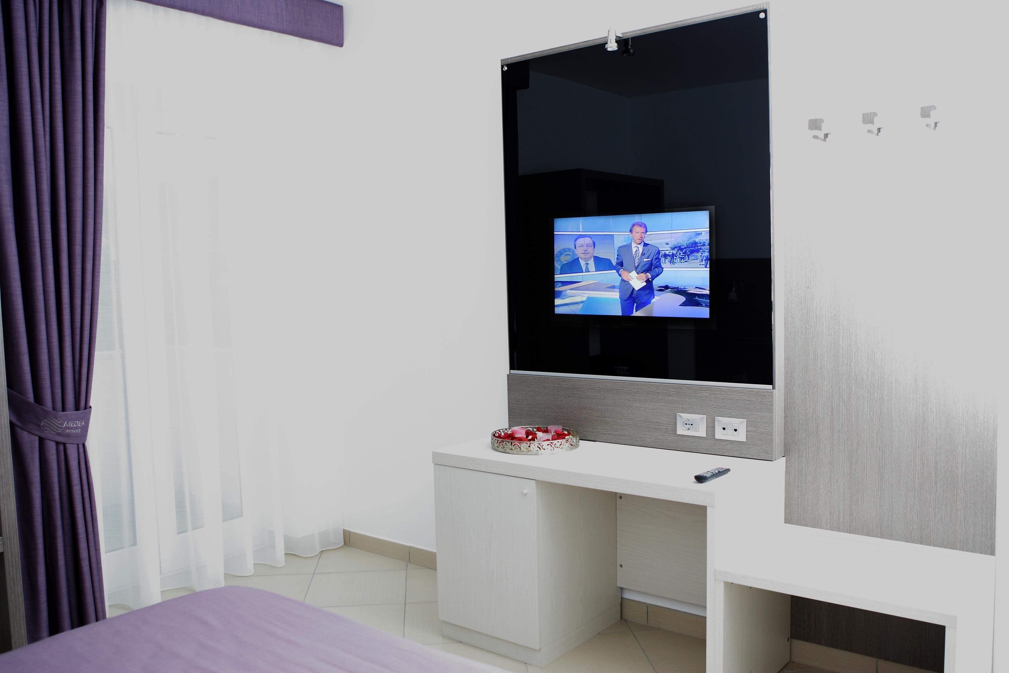 Habitación de hotel con un televisor de pantalla plana grande montado en la pared, un pequeño escritorio blanco y una cortina morada.