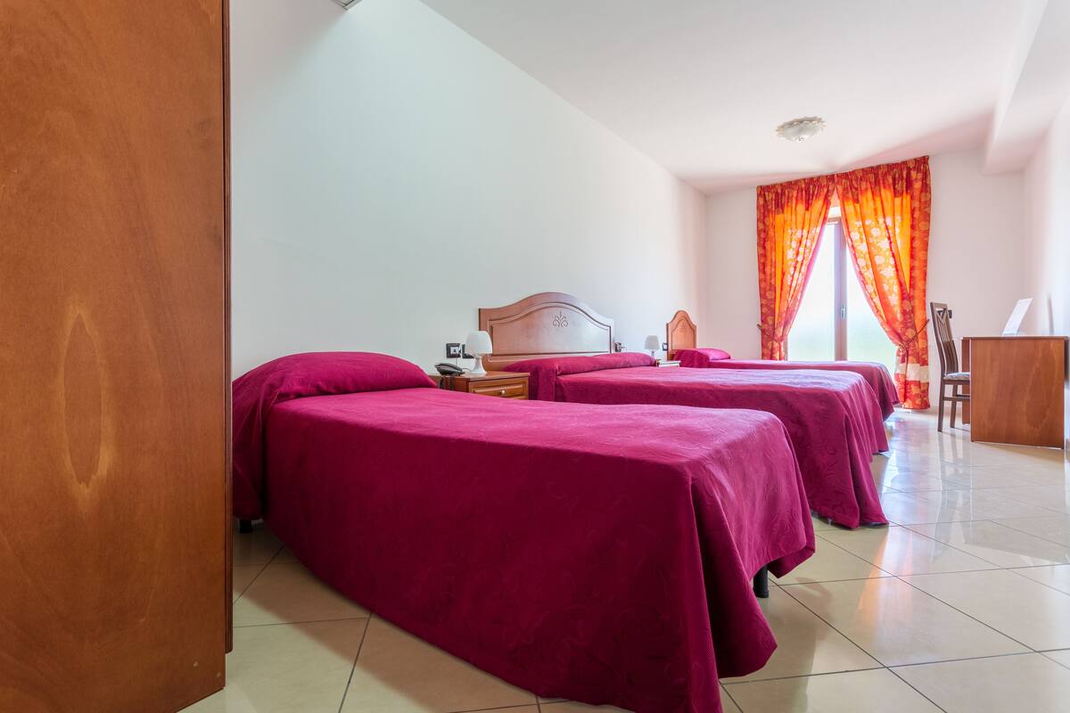 A hotel room with two beds covered in red linens, a wooden headboard, a nightstand, and a desk with a chair.