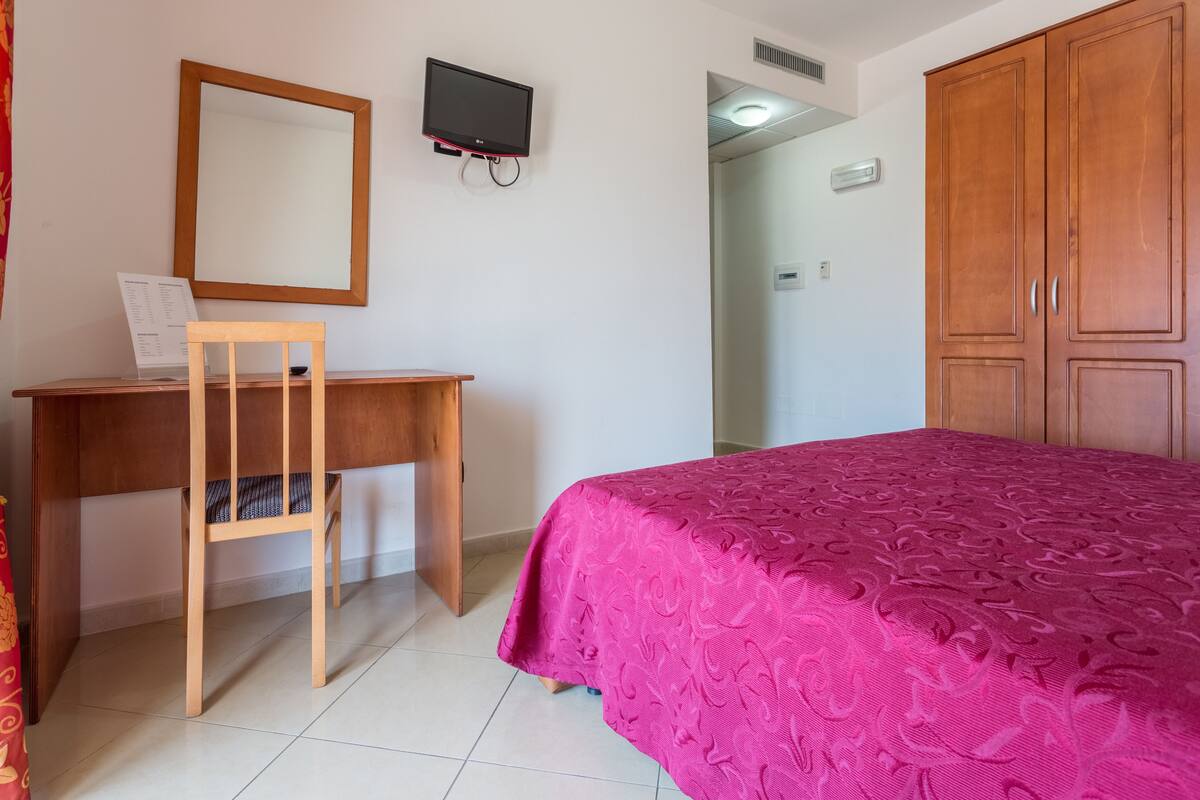 A hotel room with a wooden desk, a computer, a TV mounted on the wall, a bed with a pink bedspread, and wooden wardrobes.