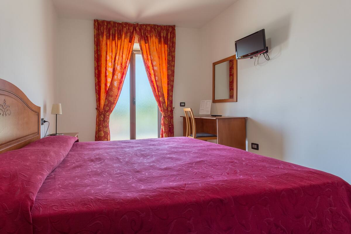 A hotel room with a large bed, a desk, a chair, a television, and a window with curtains.