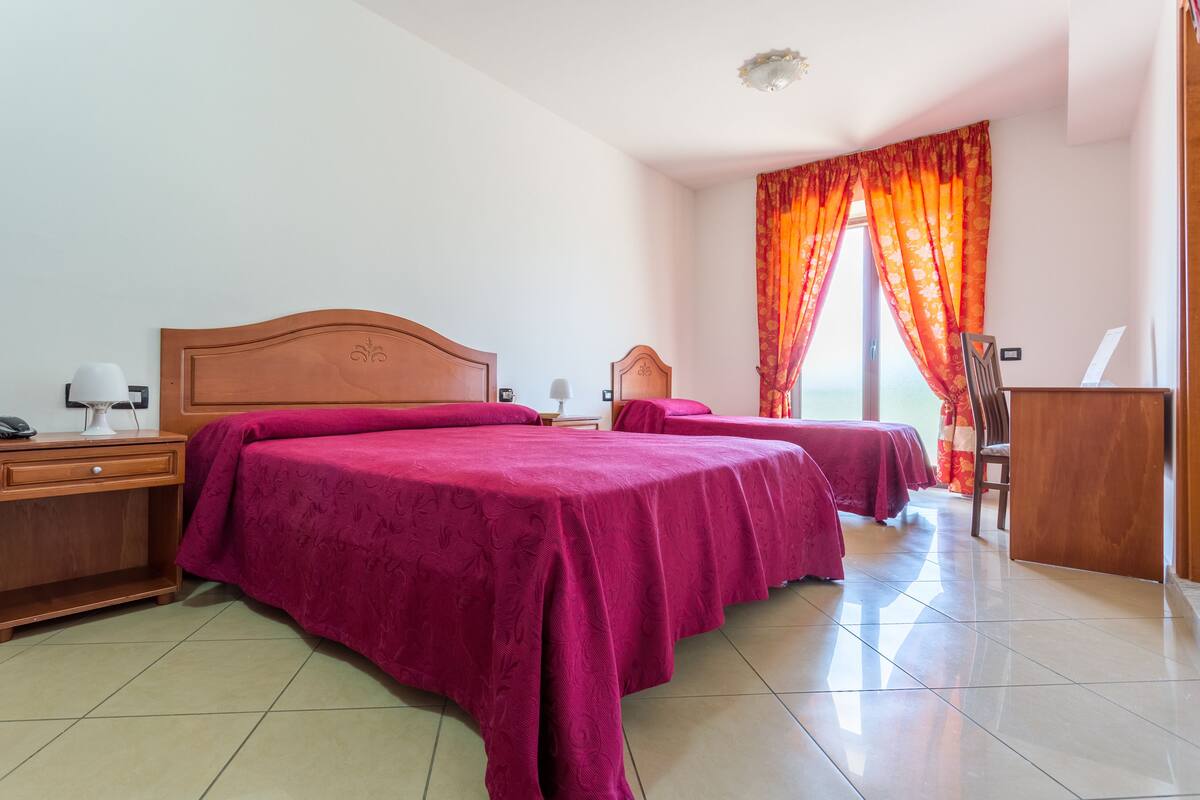A hotel room with two beds, a wooden headboard, a desk with a lamp, and a view of the sea through curtains.