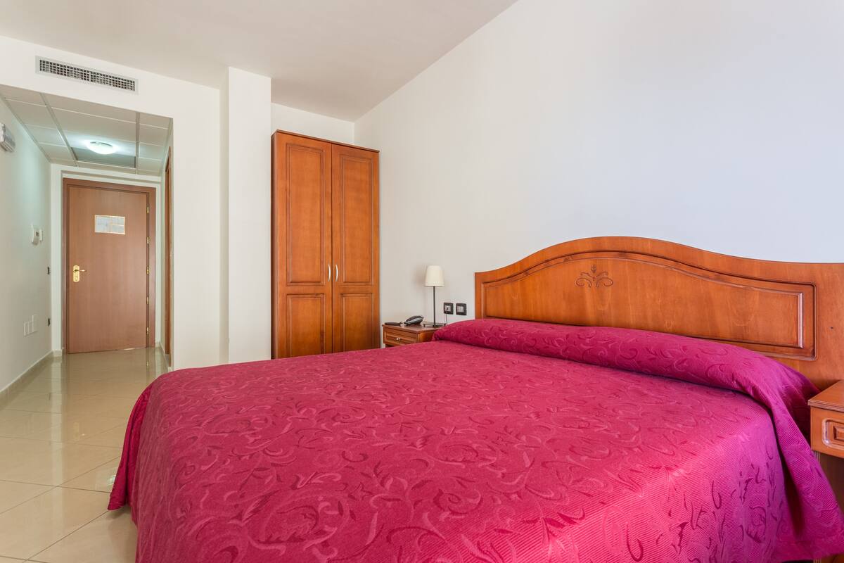 A hotel room with a large bed, wooden wardrobe, and a nightstand with a lamp.
