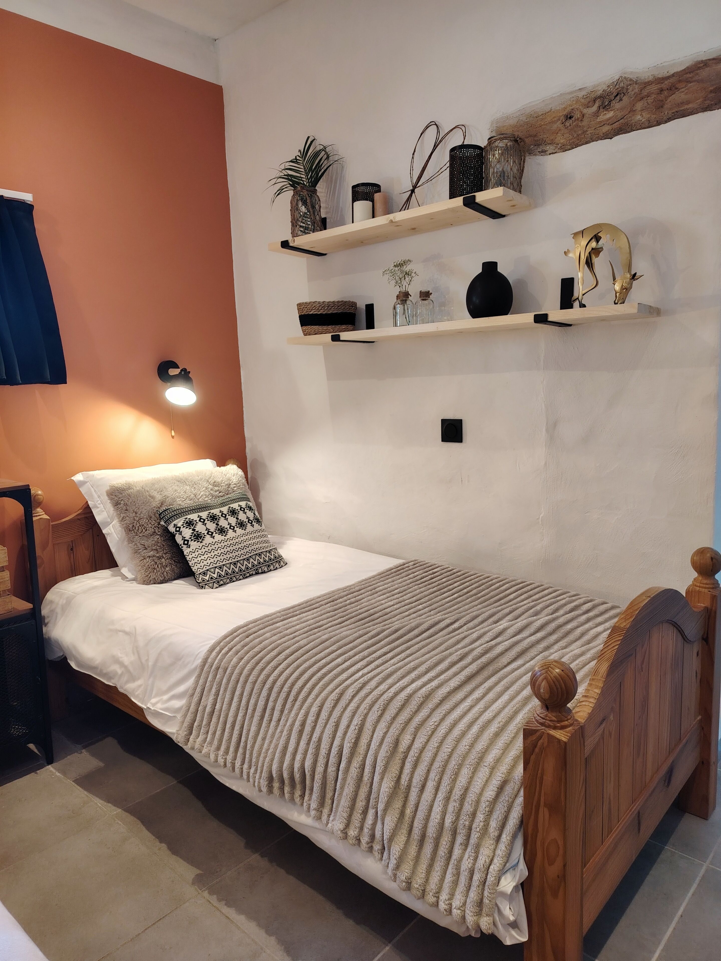 Una cama individual con cabecera de madera, una mesita de noche, un estante de pared con objetos decorativos y una lámpara de pared.