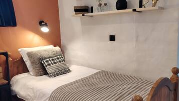 Una cama individual con cabecera de madera, una mesita de noche, un estante de pared con objetos decorativos y una lámpara de pared.
