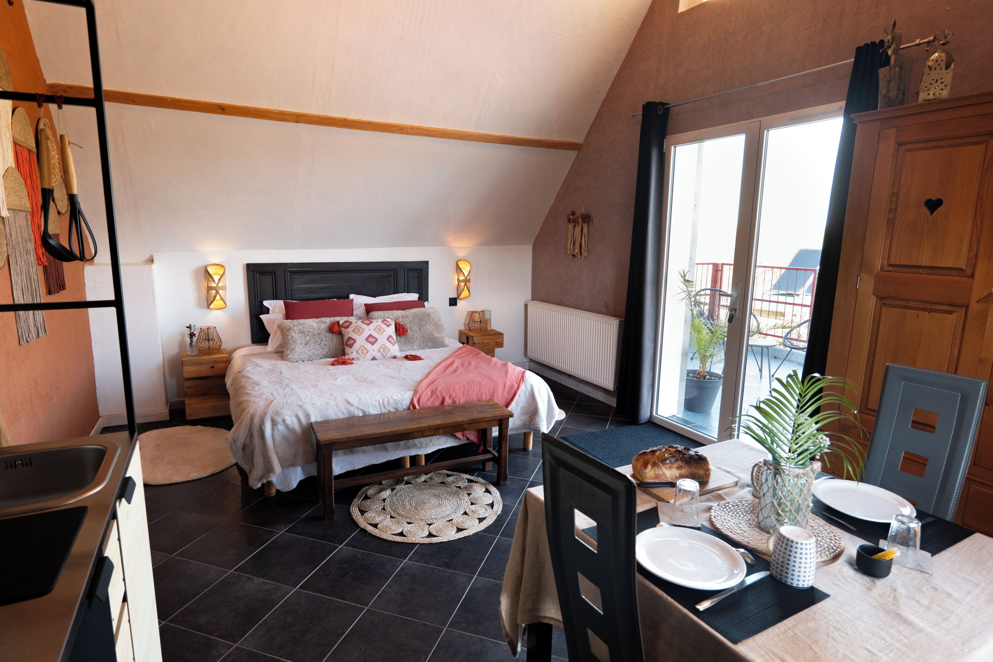 A bedroom with a bed, bedside tables, a bench, a dining table with chairs, and a view of the outside.