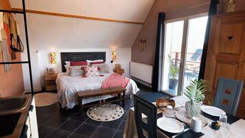 A bedroom with a bed, bedside tables, a bench, a dining table with chairs, and a view of the outside.