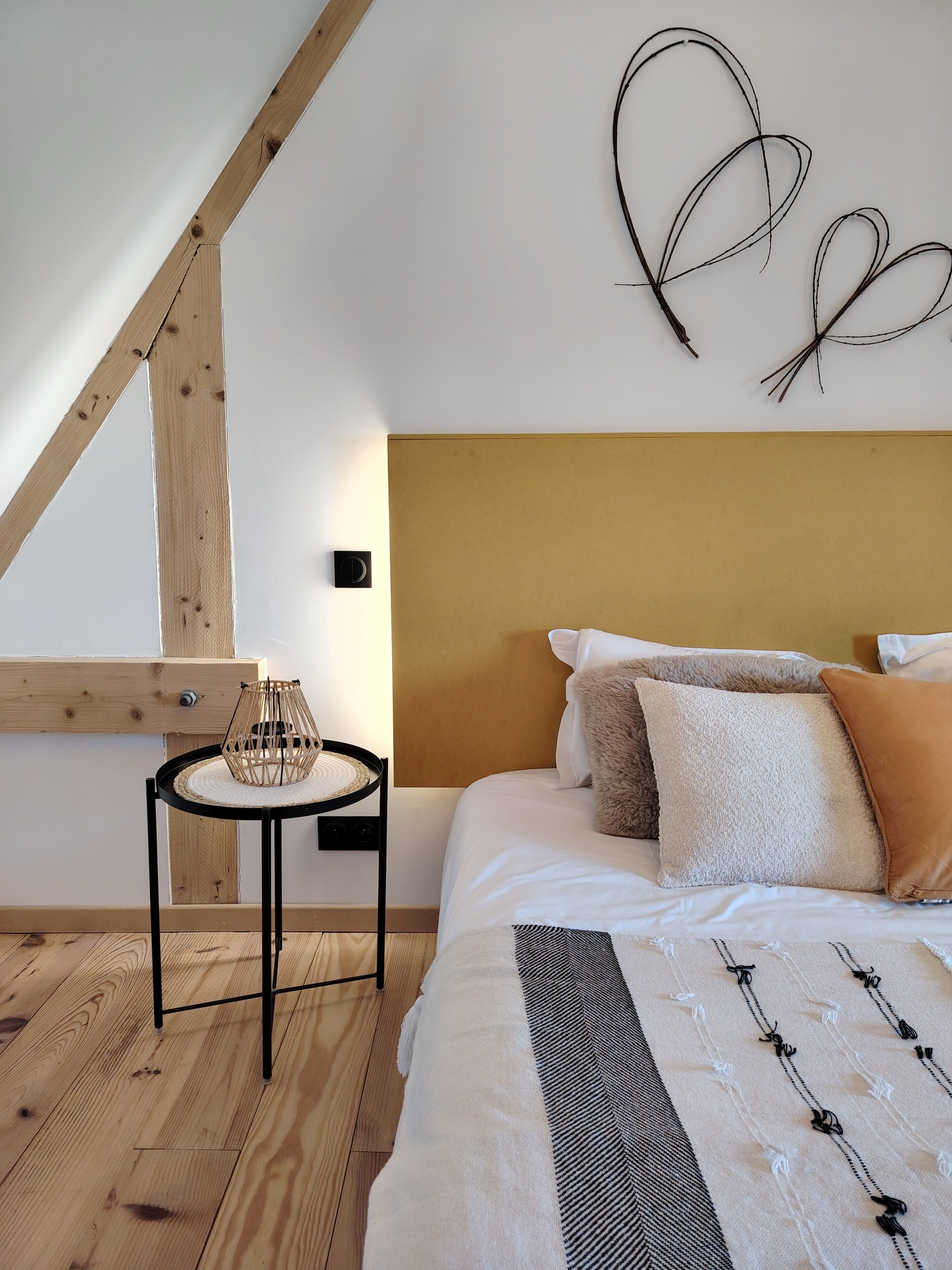 Un dormitorio con techo inclinado, una cama con muchos cojines, una mesita redonda con una lámpara de fibras naturales y una obra de arte abstracto colgada en la pared.